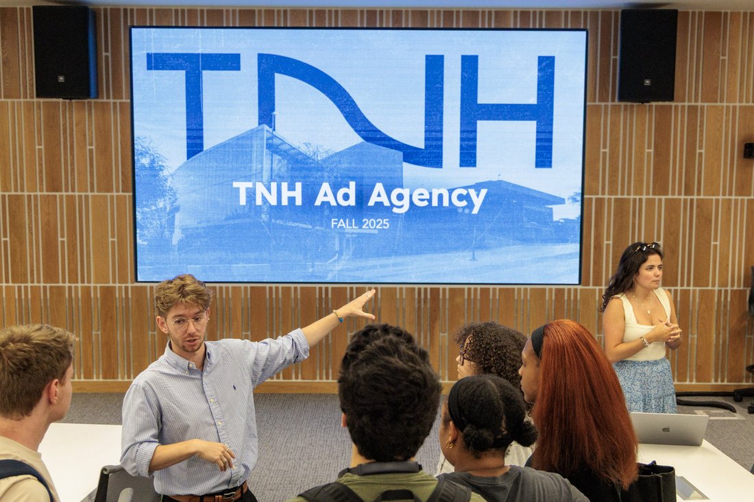 A student gestures toward a screen reading "TNH Ad Agency, Fall 2025" while presenting to classmates during an ad agency meeting..