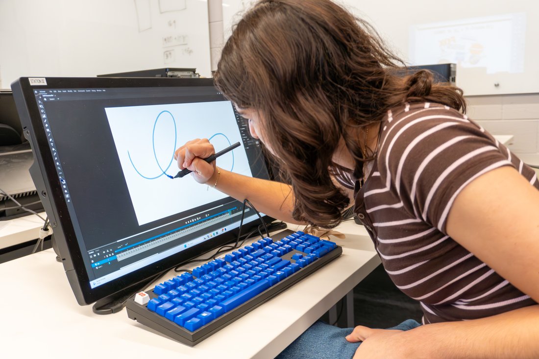 A student sits at a computer workstation and draws circular lines on the screen with a stylus.