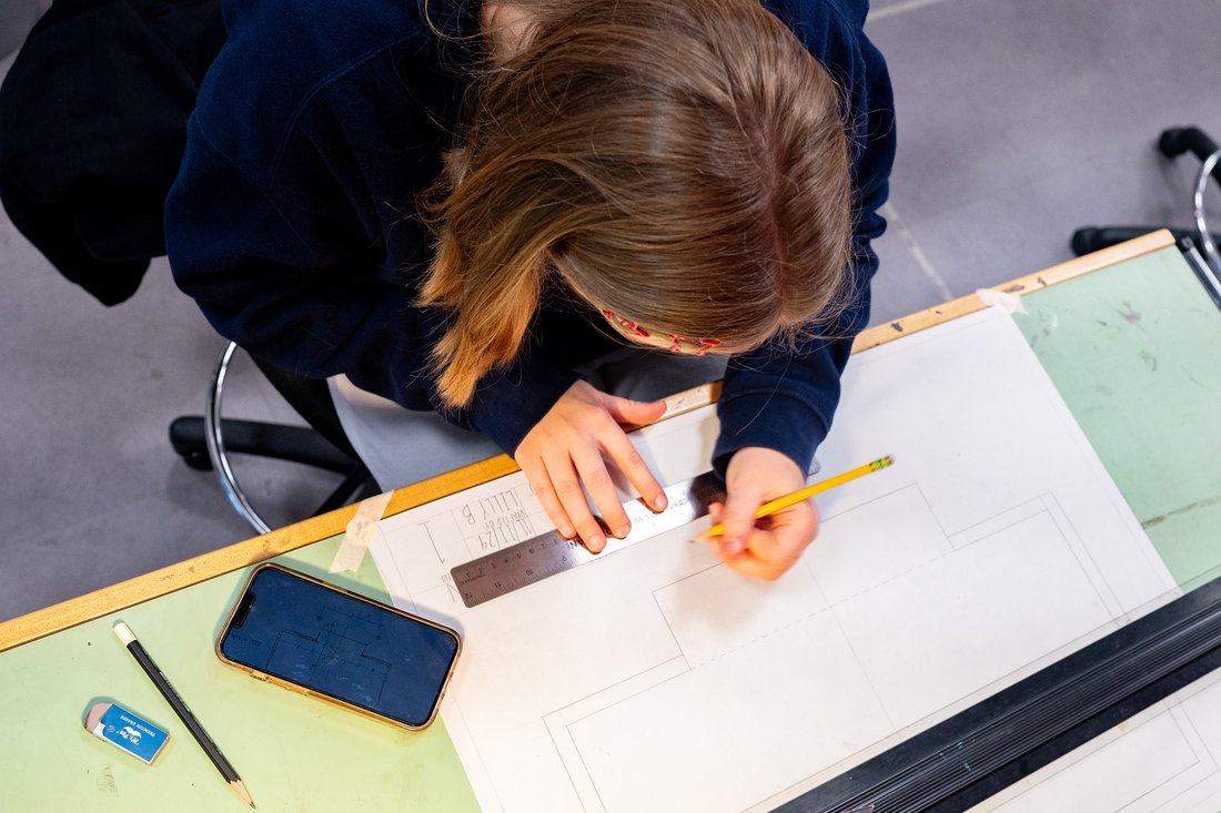 A person leans over a drafting table and draws the basic lines of a stage with a pencil.