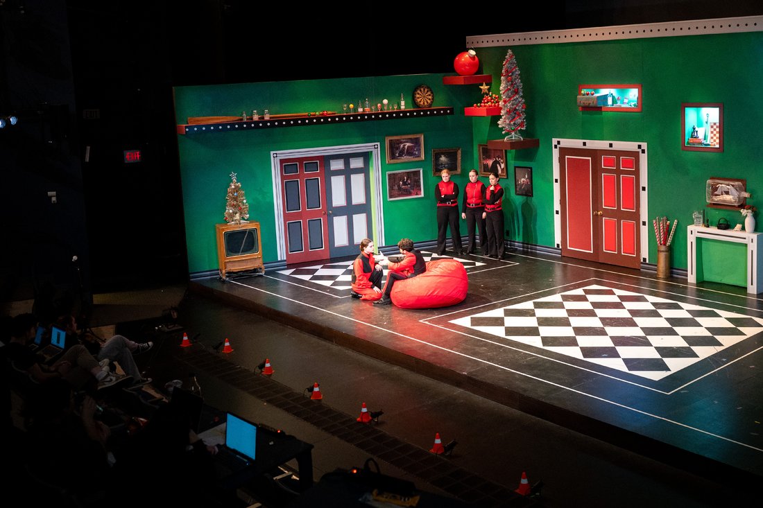A wide shot of a theater stage with actors in costume on a set that has a checkerboard floor and bright colors on the walls.
