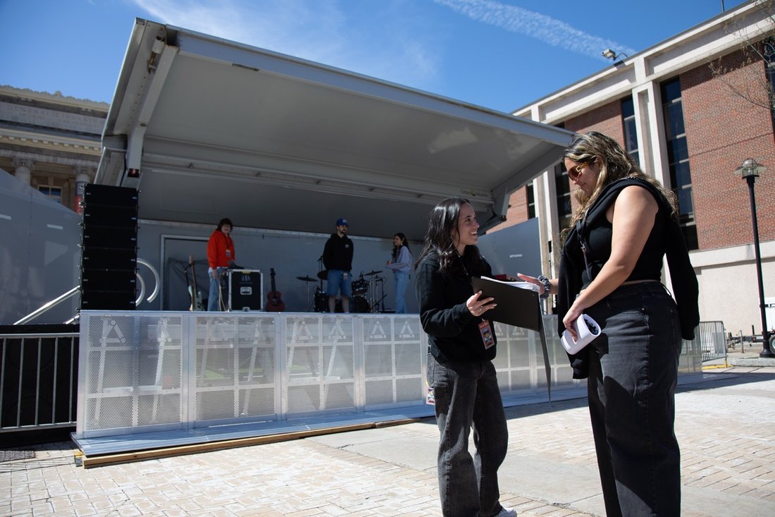 Two Bandier students talk in front of a stage at Block Darty 2024. On the stage, students set up equipment.