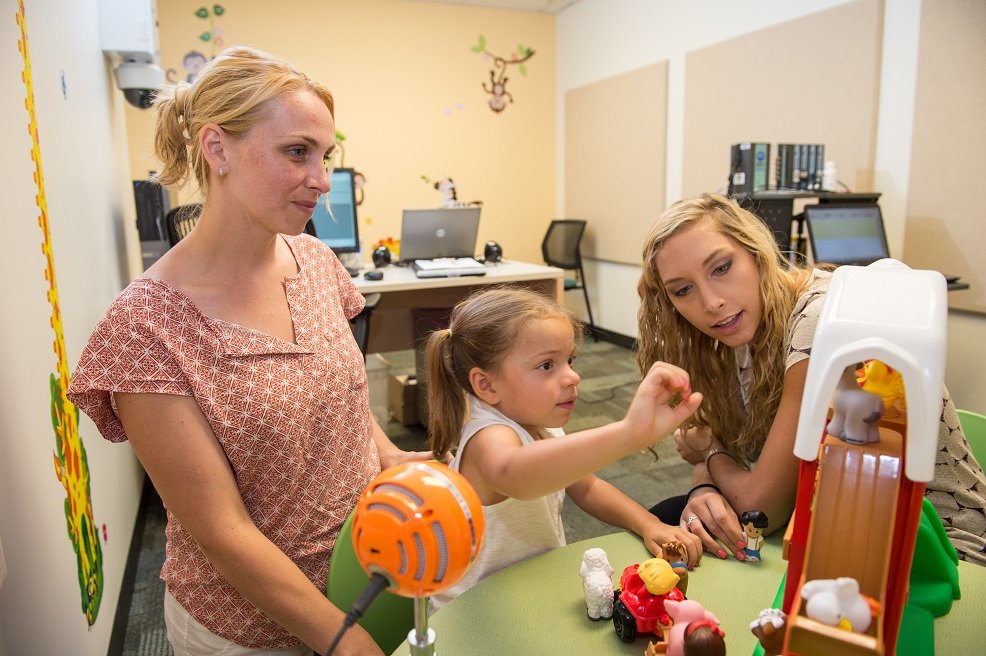 A child holding a toy while two adults watch.