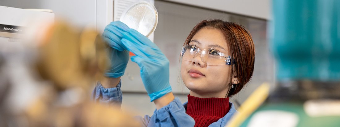 A person wearing a lab coat and safety glasses holding a lab dish