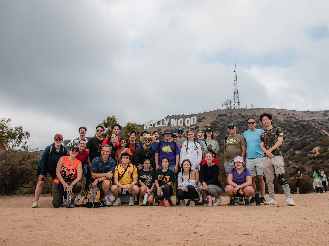 A group of people pose and smile on a trail with a large Hollywood sign in the hills behind them.