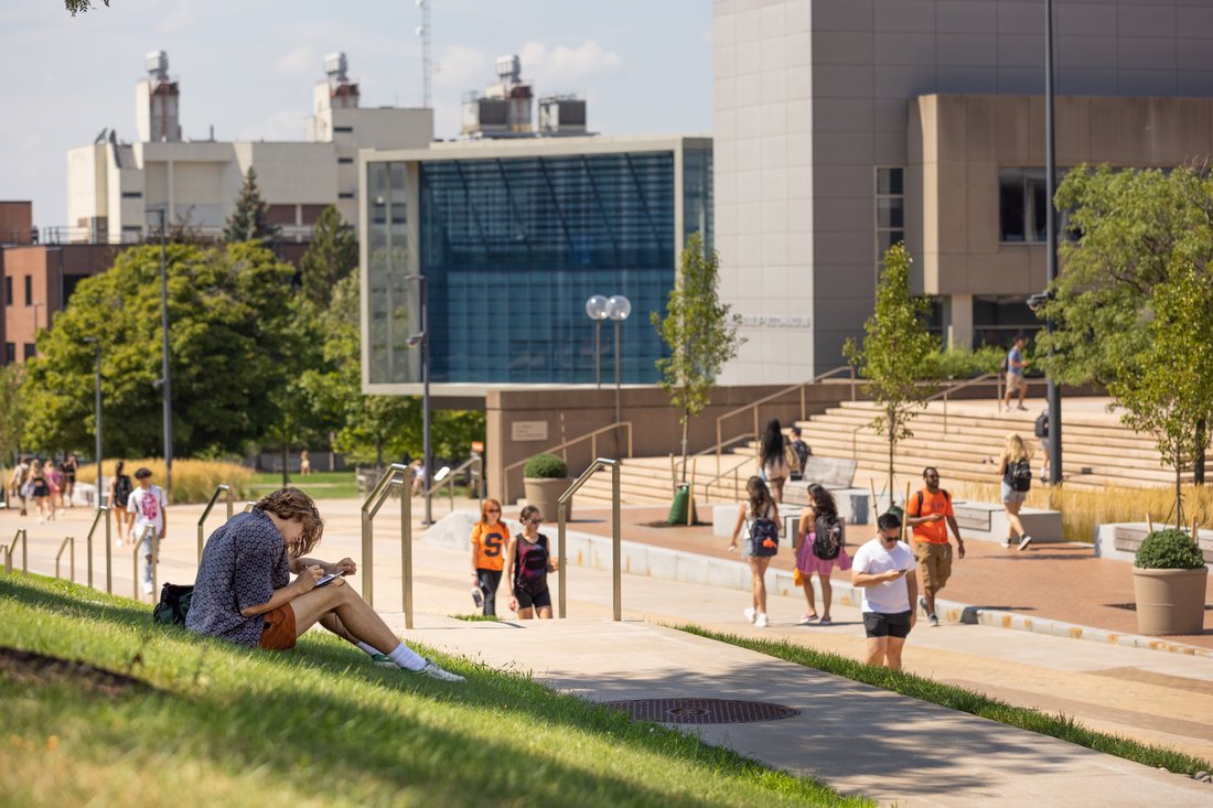 A student sitting in the center of campus, working on a writing project. The Newhouse School and other campus buildings can be seen in the background.