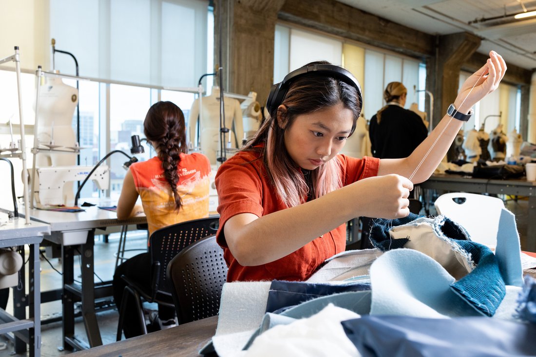 A person sits at a work table sewing fabric. Behind her is another person working at a sewing machine.