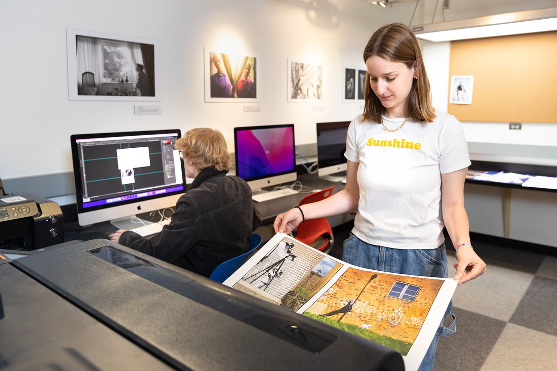 A woman looks at large color photos coming out of a printer while a man works on a computer in the background.