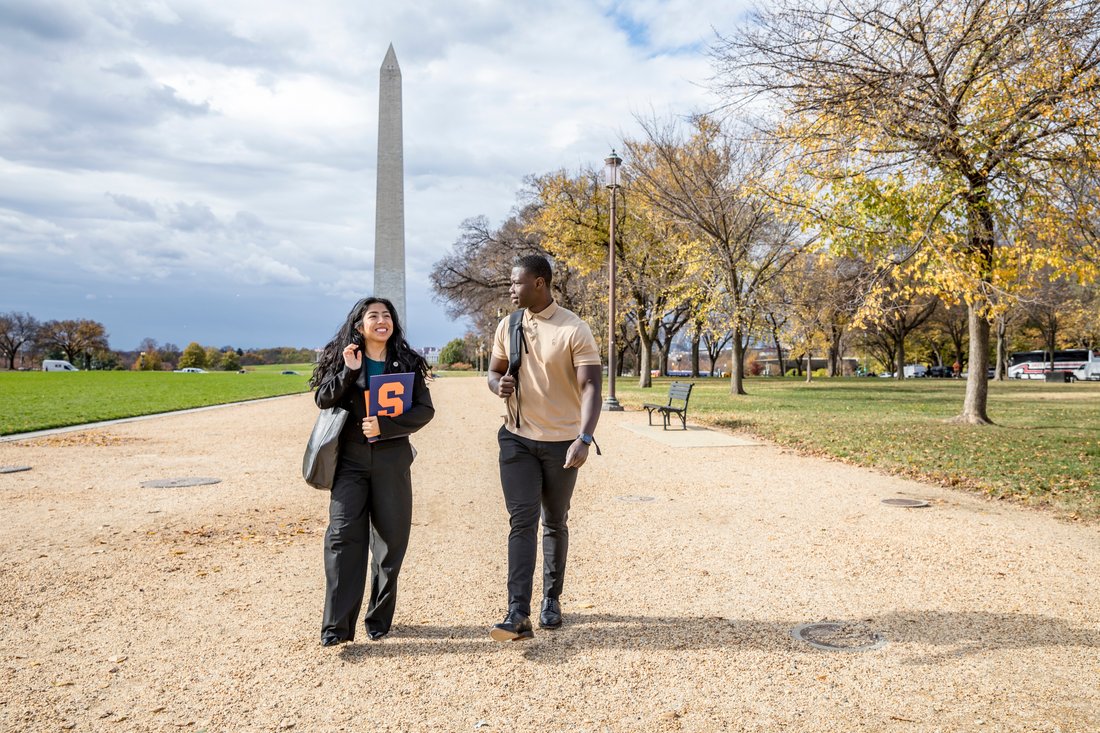 Two people carrying a Syracuse folder walk on a path near the Washington Monument in fall.