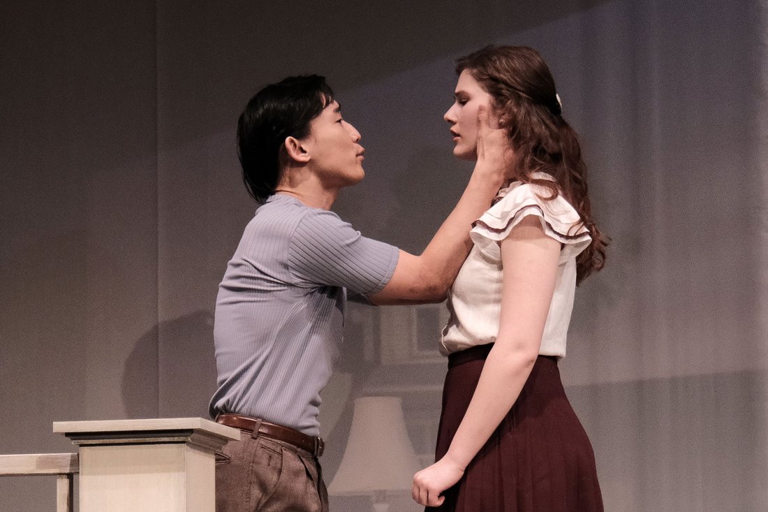 Two actors in costume on a stage face each other, with one actor cupping the other actor