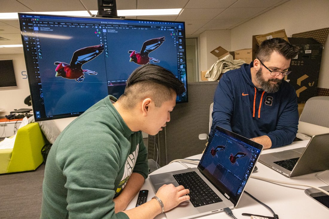 A student and a professor each look at their computers at a table as large monitor in the background shows drafts of a computer illustraiton of a red bird