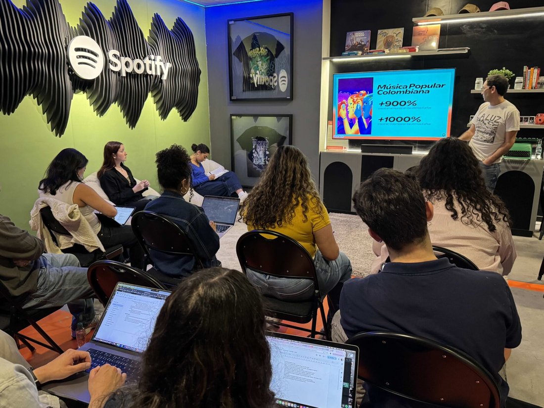Students with computers on their laps sitting in metal chairs face an instructor looking at video screen in conference room with the "Spotify" logo on the wall.